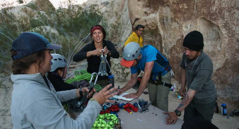 A group of people stand around a table set up outside. There appears to be climbing gear scattered on it. 